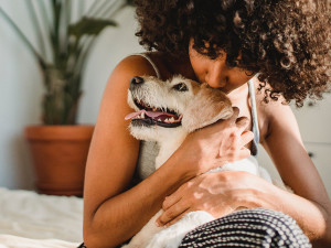 A woman sitting on a bed holding a dog close in her arms. 