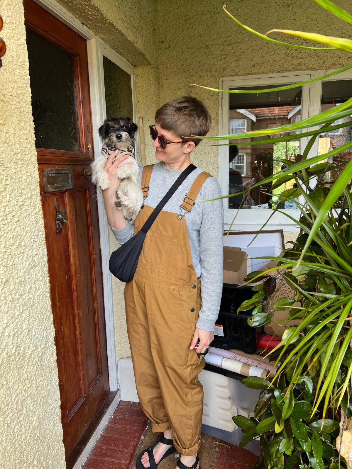 A woman in dungarees with short hair holds up a small dog next to a front door