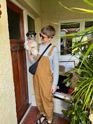 A woman in dungarees with short hair holds up a small dog next to a front door