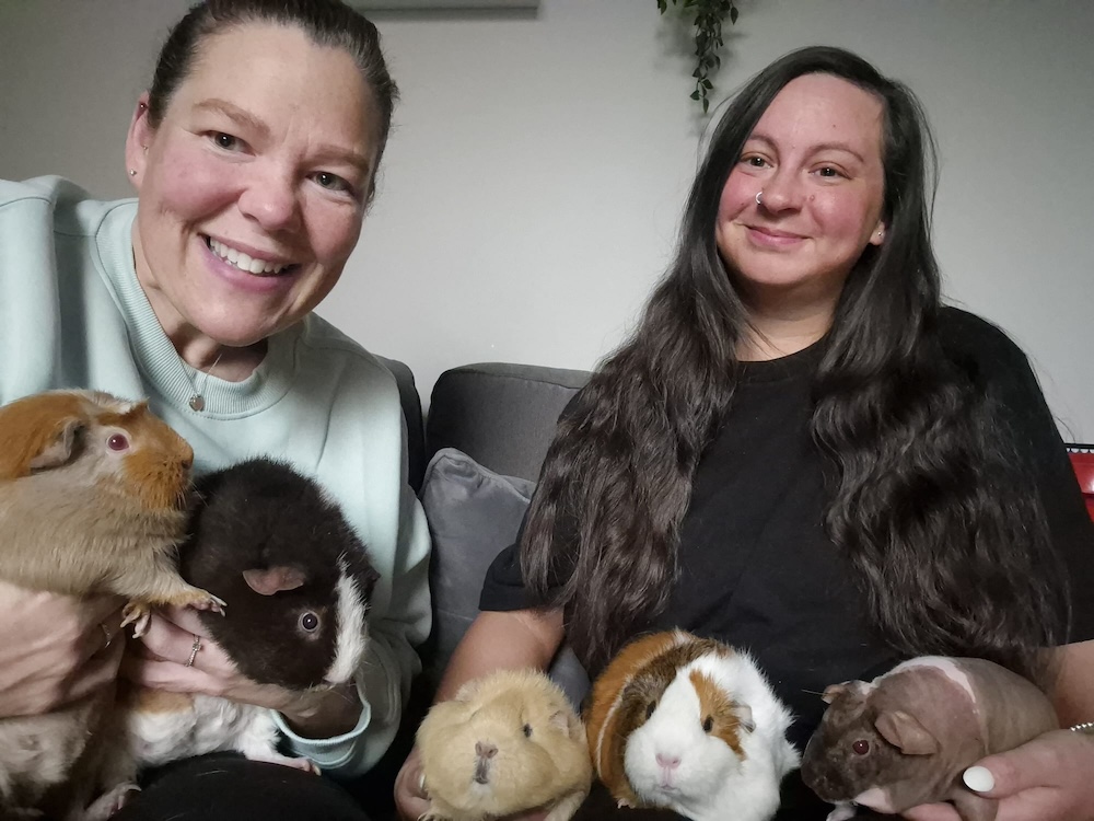 two women with brown hair hold five guinea pigs and smile at the camera