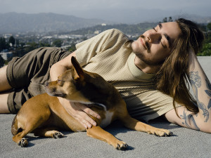 Tyler Gaca basking in the sun laying on concrete while holding his Chihuahua dog against a mountainous backdrop