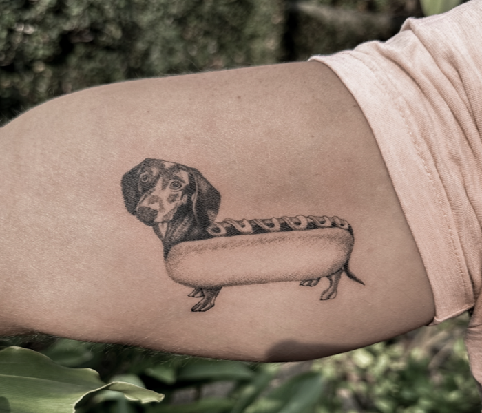 A black and grey tattoo of a Dachshund in a hot dog bun. 