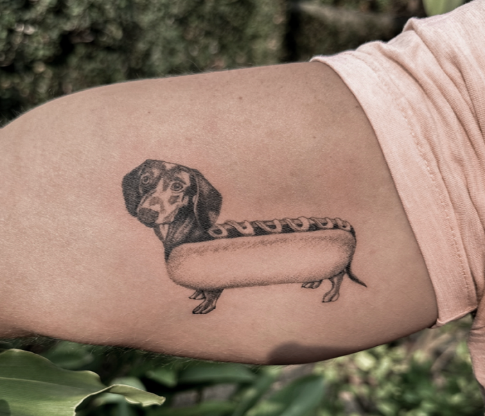 A black and grey tattoo of a Dachshund in a hot dog bun.