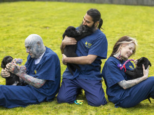 A man with a face tattoo, a man with a beard and a woman with face tattoos hug black labrador puppies