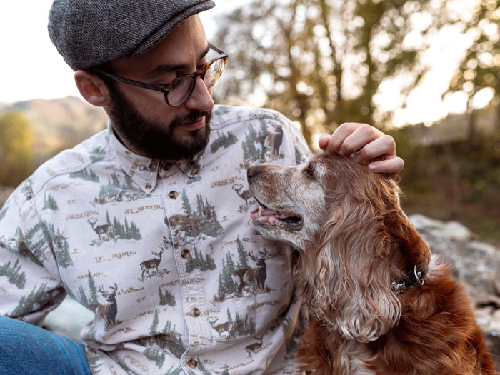 Man with beard and wearing glasses and a grey hat sitting outside petting the head of his old foster dog