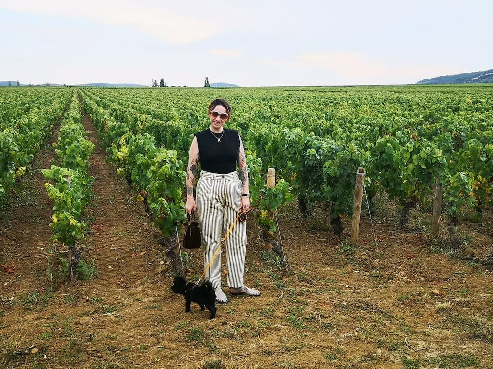 a woman stands with her small black dog in a vineyard