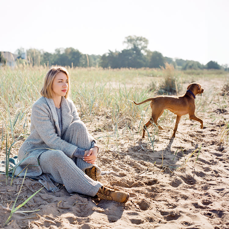 A woman with blonde hair sits on beach while her dog trots off on the sand