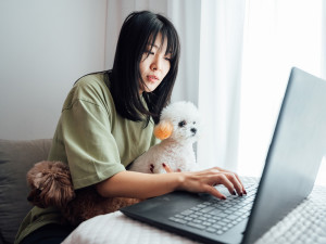A dark-haired woman holding her two poodle mix dogs, one white and one brown, while working on her computer.