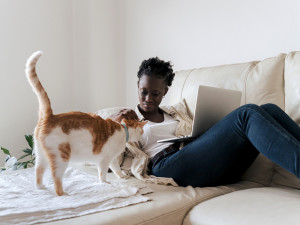 Black woman sitting on her couch with laptop and petting her cat 