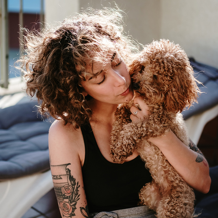 Curly haired woman with tattoos holding her Labradoodle dog up to her face in a tender embrace