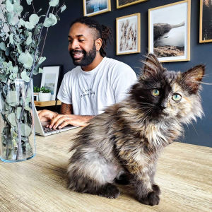 A man sitting at a table with his laptop and cat. 