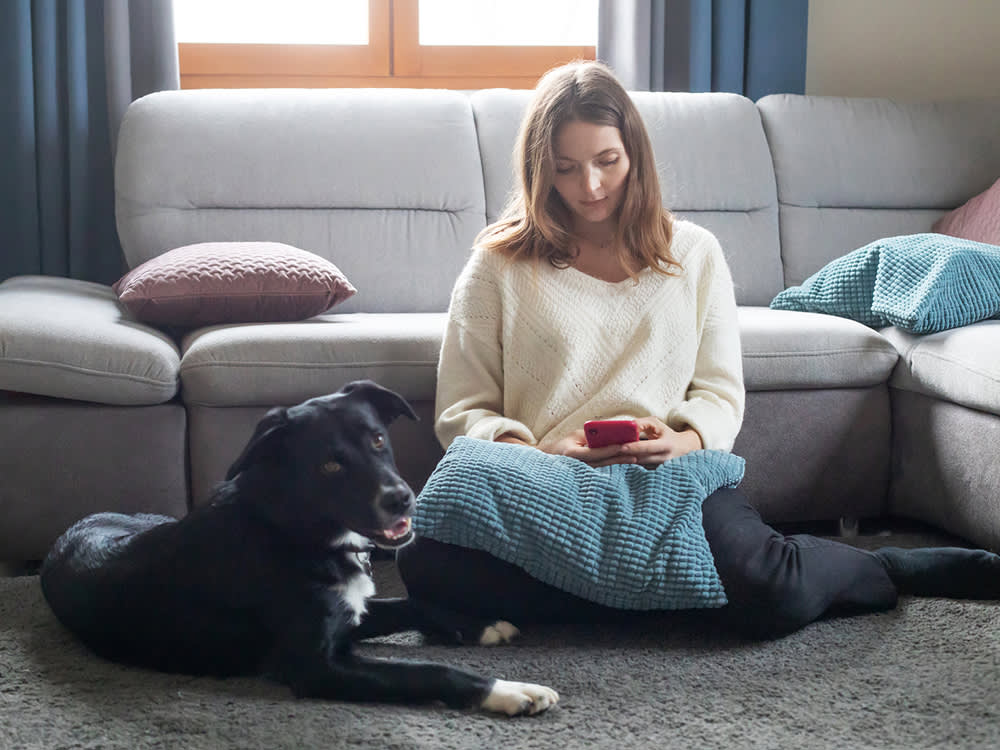 A woman with shoulder length hair wearing a white sweater and black leggings sitting on the floor in front of her couch next to a black dog with white spots while looking at her phone 