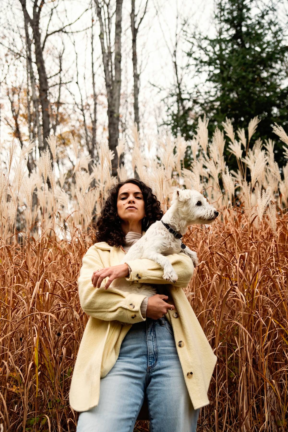 a woman with curly black hair holds a small white dog and scowls down at the camera