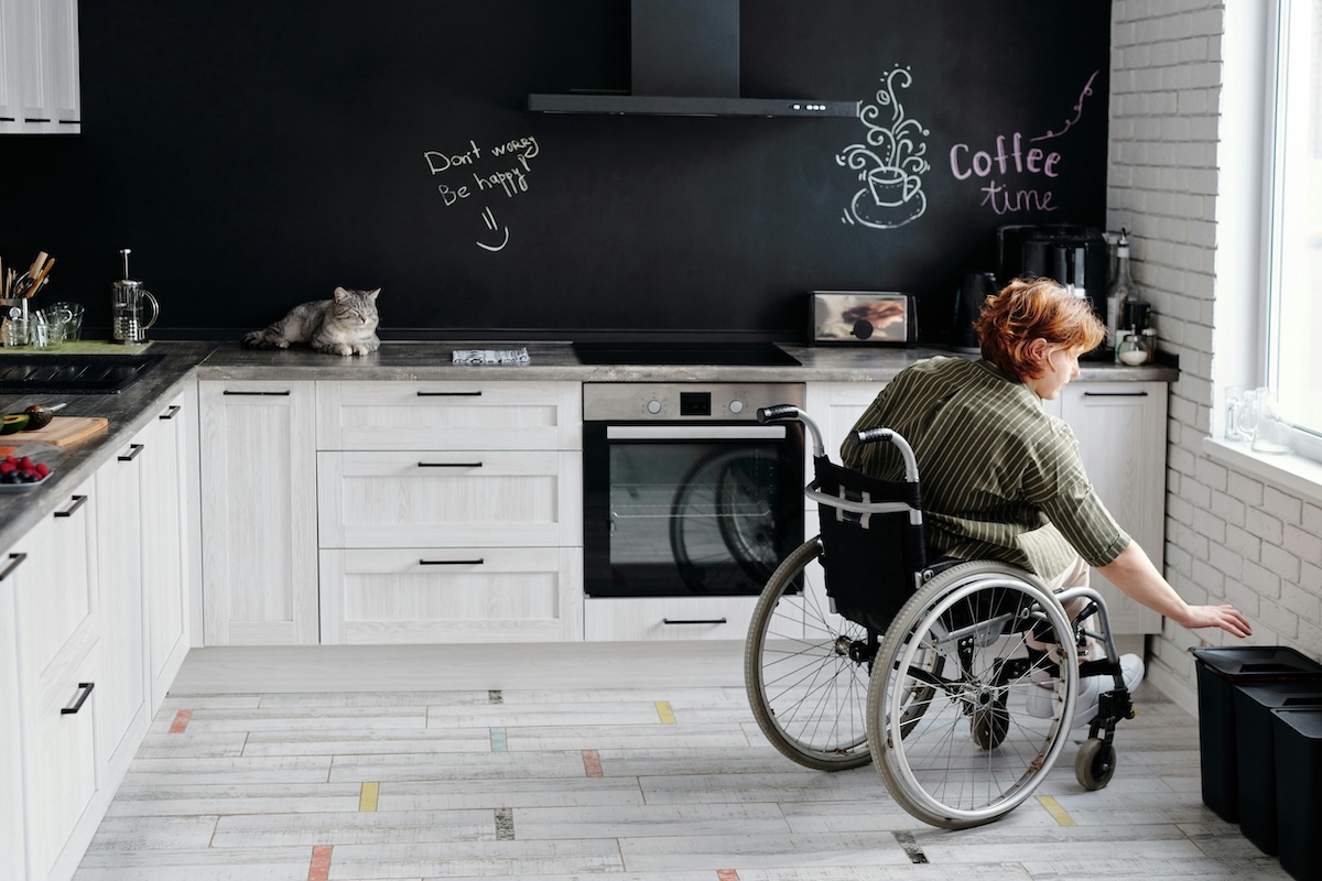 a woman in a wheelchair puts something in the bin in her kitchen, a grey cat sits on the counter