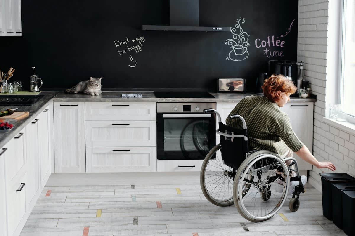 a woman in a wheelchair puts something in the bin in her kitchen, a grey cat sits on the counter