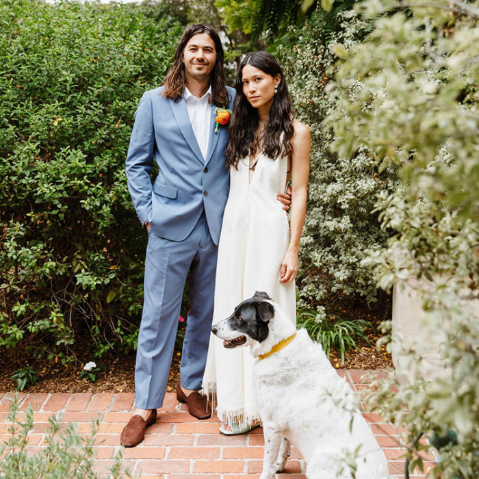 Calley Benoit Belli  wearing a wedding gown standing next to her husband who is wearing a light blue suit with their dog sitting on the red brick walkway in front of them