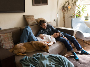 A couple laying on their couch in a cozy looking living room with their large mixed breed foster dog 