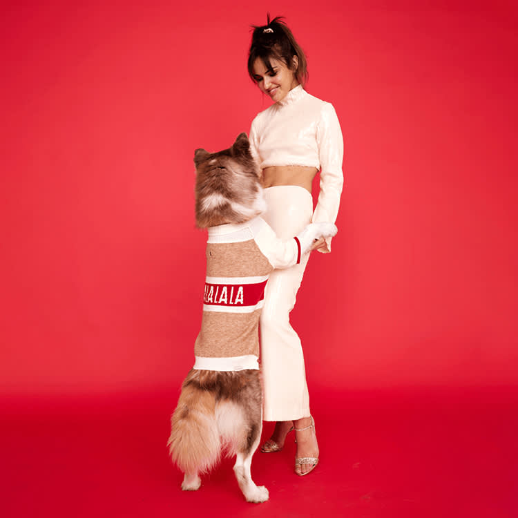 Woman in a sweater standing and holding the two front legs of a Husky dog wearing a matching sweater on a red background