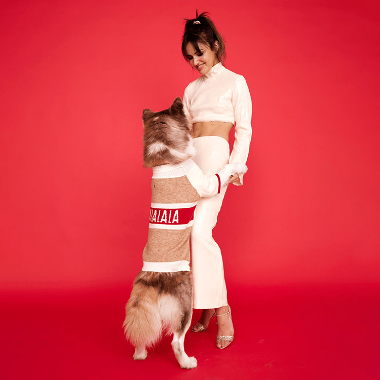 Woman in a sweater standing and holding the two front legs of a Husky dog wearing a matching sweater on a red background