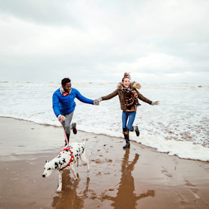 A man and woman on the beach running out of the waves wearing coats and boats and holding a lead with a Dalmatian on the end