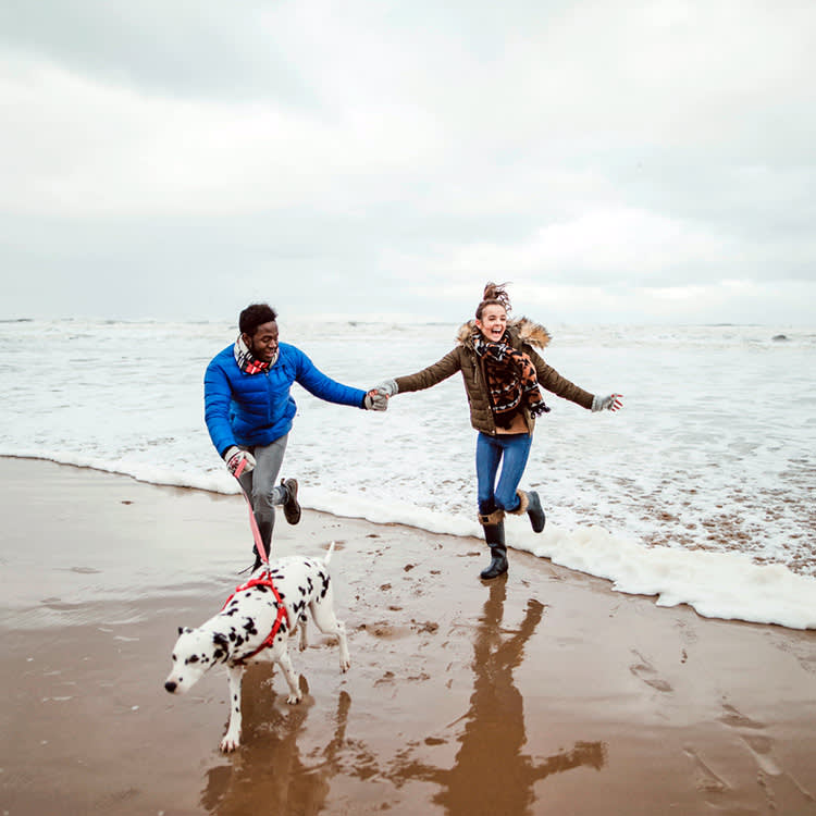 A man and woman on the beach running out of the waves wearing coats and boats and holding a lead with a Dalmatian on the end