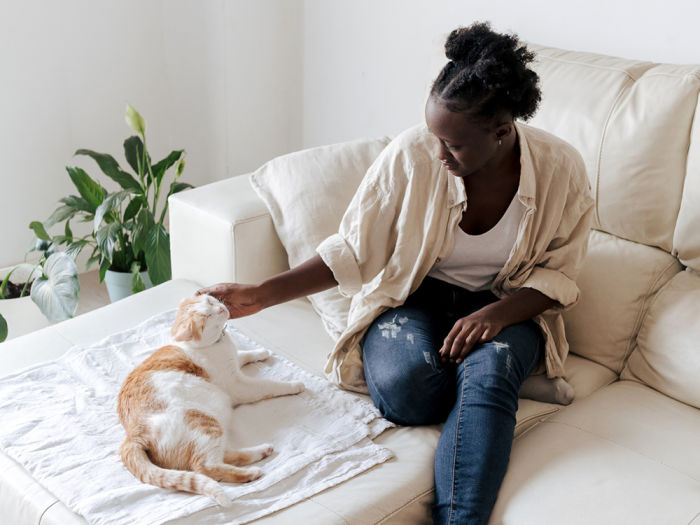 Woman with tight curly black hair wearing a tan linen shirt and jeans sitting on a cream couch petting her tan and white cat who is laying on a blanket on the couch int he living room