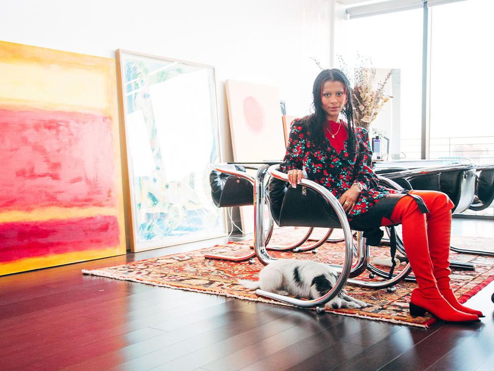 Sami Miró sitting on a black chair with her small white rescue dog under the chair