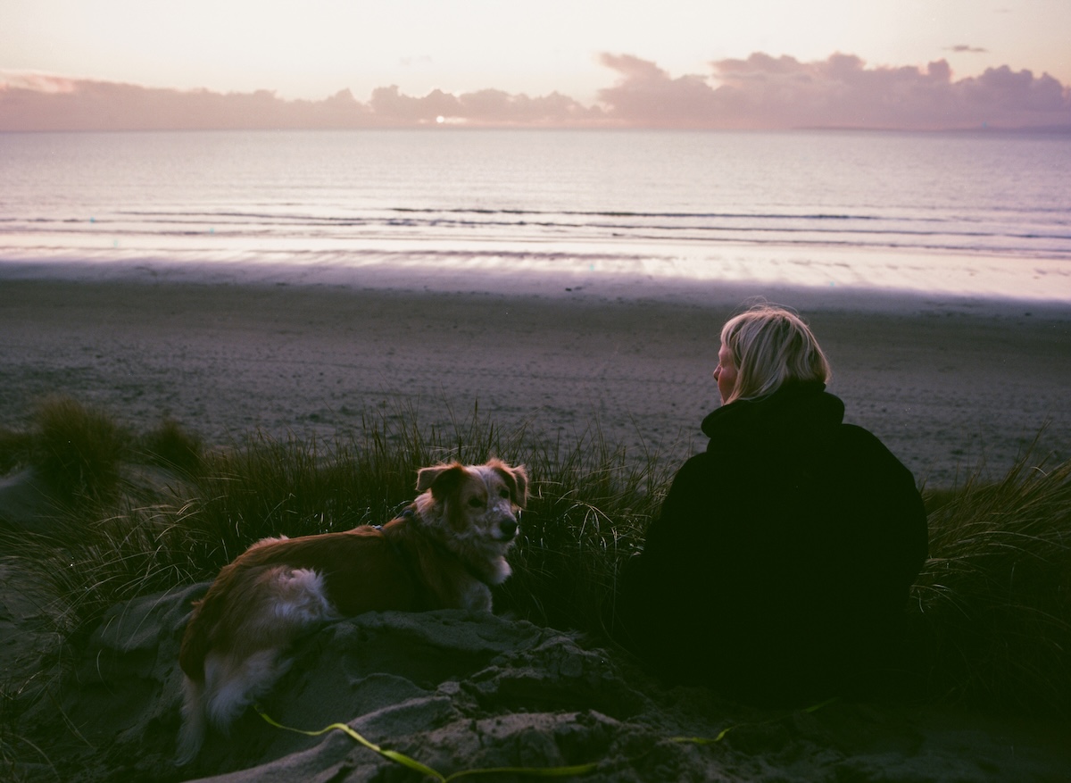 a woman and a dog sitting on a beach at sunset in winter
