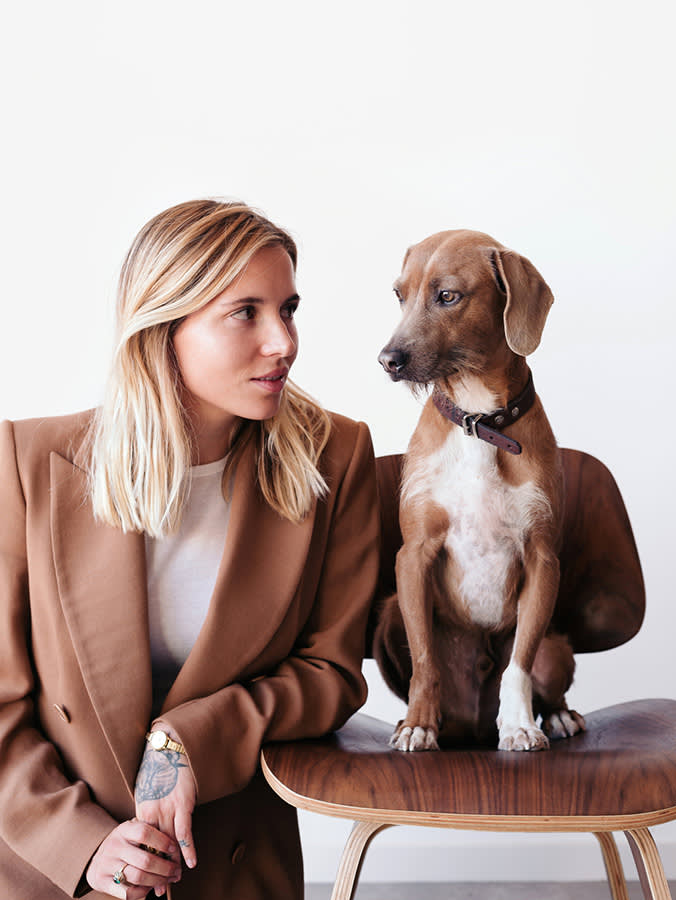 Brown, half breed dog sitting on a chair next to his owner, a blonde stylish woman wearing a brown smart suit.