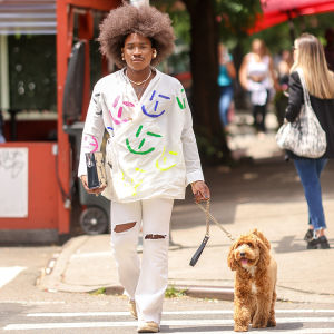 Person with a large curly brown afro wearing a stylish two-piece white suit with multicolors designs on the coat walking their dog in the city