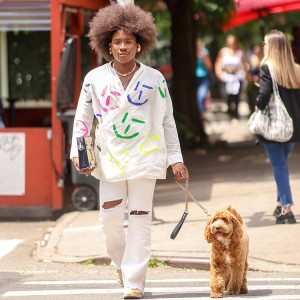 Person with a large curly brown afro wearing a stylish two-piece white suit with multicolors designs on the coat walking their dog in the city