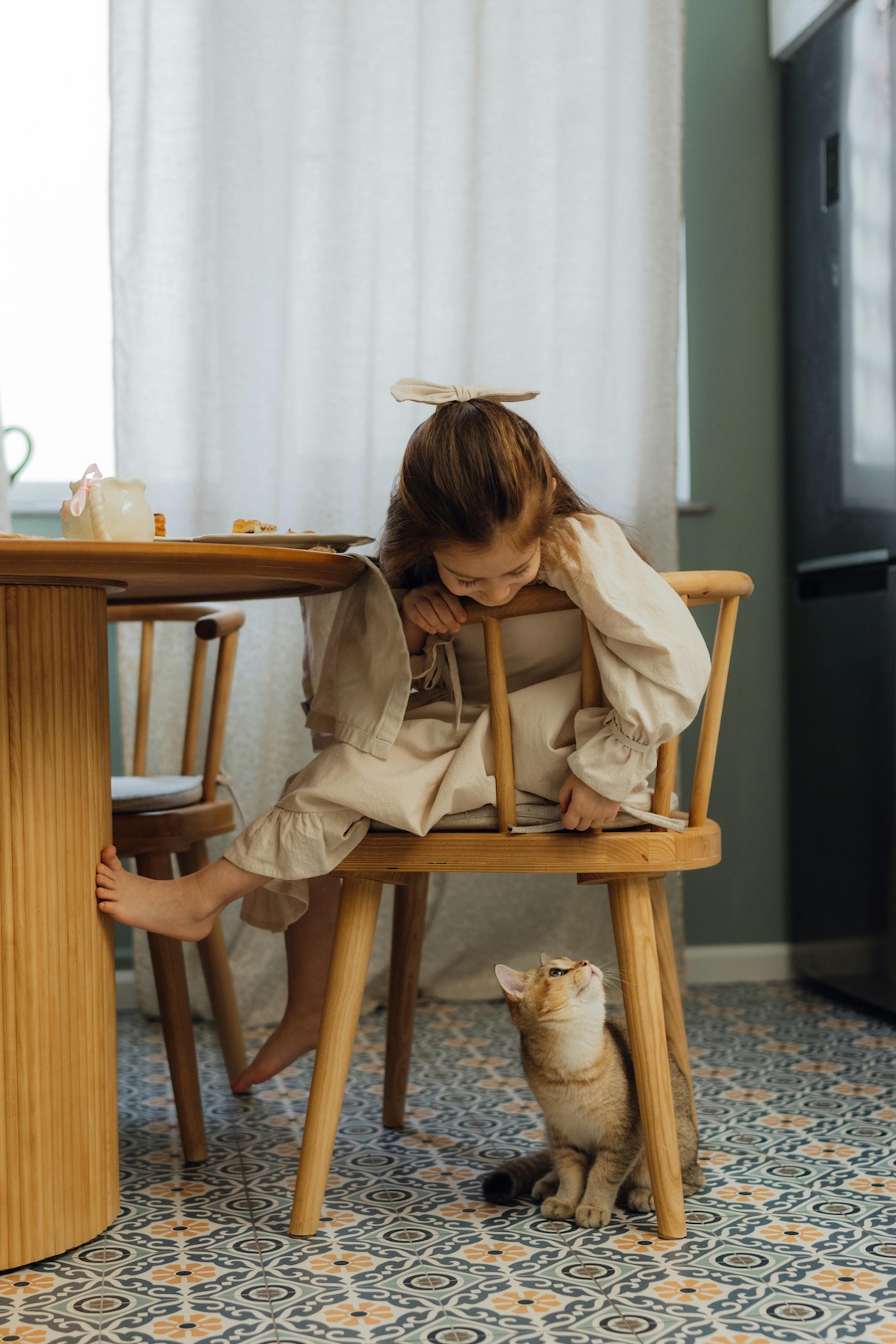 picture of a cat under a chair looking up at a little girl sitting on the chair