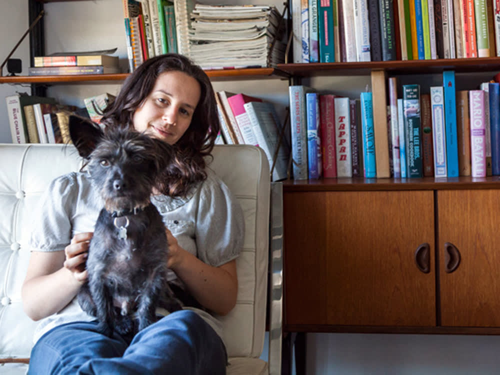 Jessica Koslow sitting on a beige couch with her black wire-haired dog sitting in her lap with a bookshelf in the background