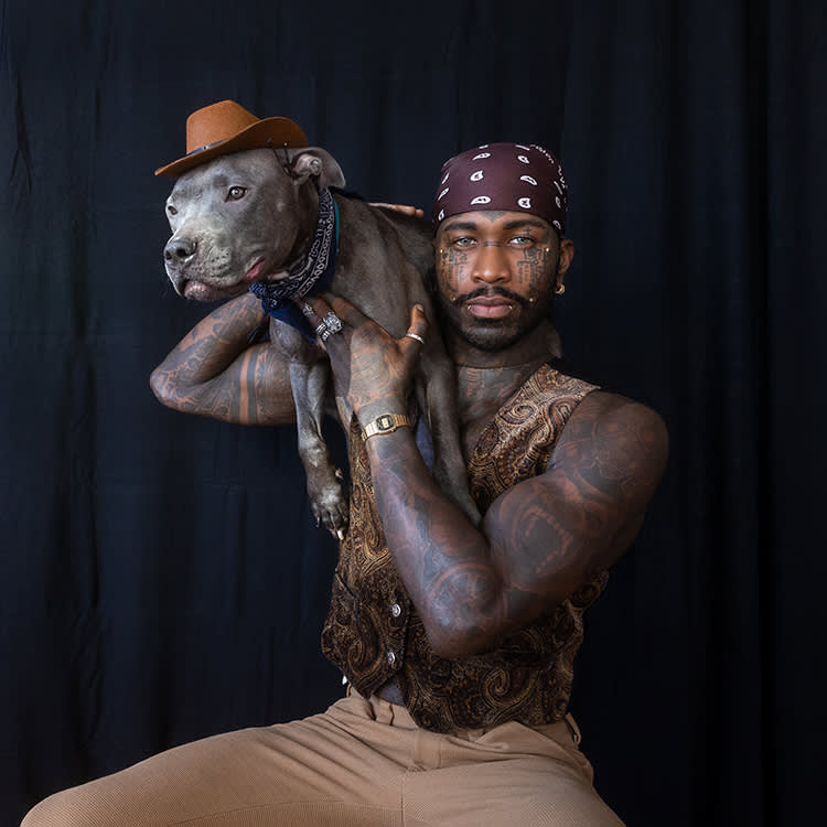 Yves, a man with a lot of tattoos and a brown paisley head bandana in tan pants holding his grey Pitbull dog who is wearing a tiny cowboy hat over his shoulder