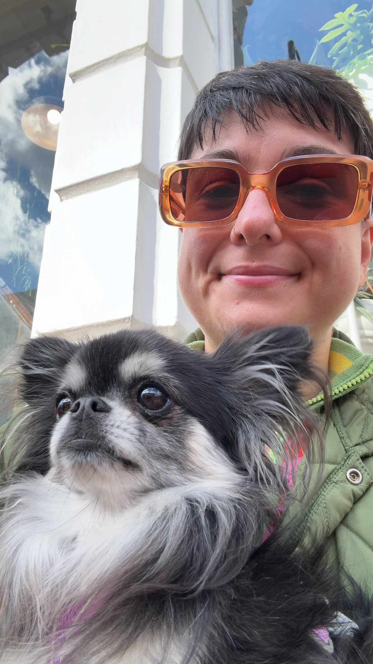 a selfie of a person with short hair wearing orange sunglasses and hugging a small fluffy dog