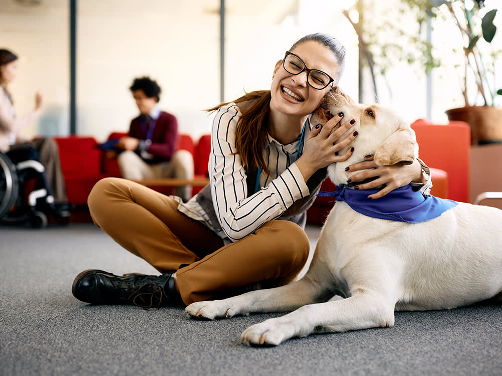 a woman in glasses smiles and sits cross-legged hugging a dog with a neckerchief