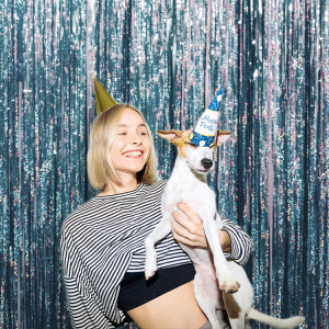 Blonde woman in a golden party hat, holding a white dog in her arms, laughing at it. The party hat on the dog slipped on it's eyes. Portrait of a pet and an owner celebrating birthday together.