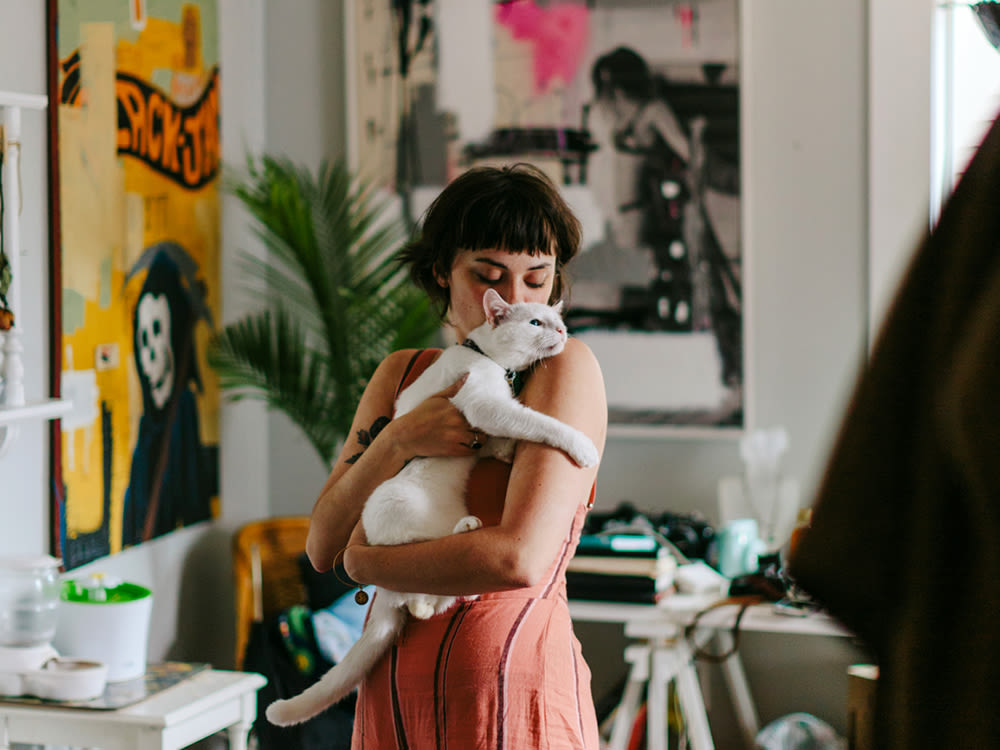 A woman with tattoos hugging her white cat in a cluttered, art-filled room
