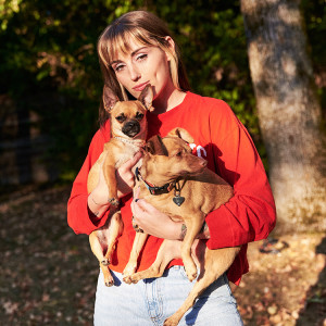 Sasha Alex Sloane wearing a bright red sweatshirt and light blue jeans holding her two small tan dogs while standing outside in the woods