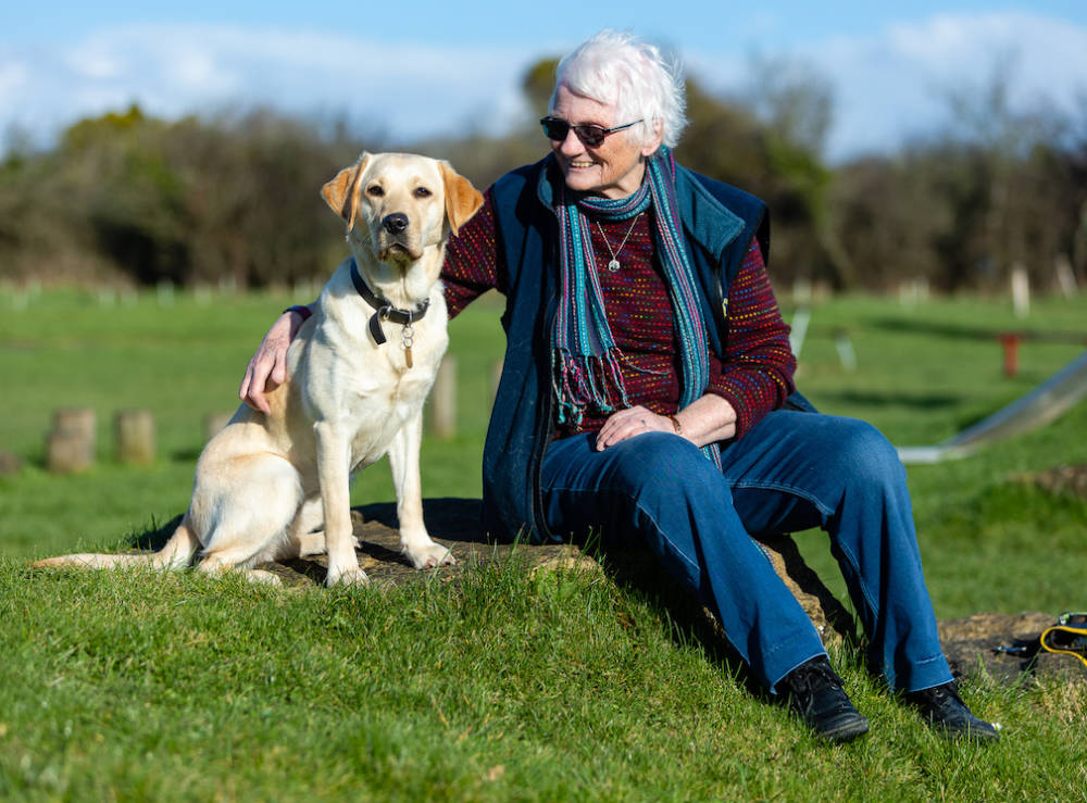 a smiling woman sits on a hill with her arm around a golden labrador