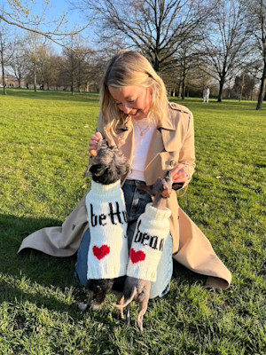 a woman with blonde hair sits in a park with two little dogs in sweaters jumping up on her