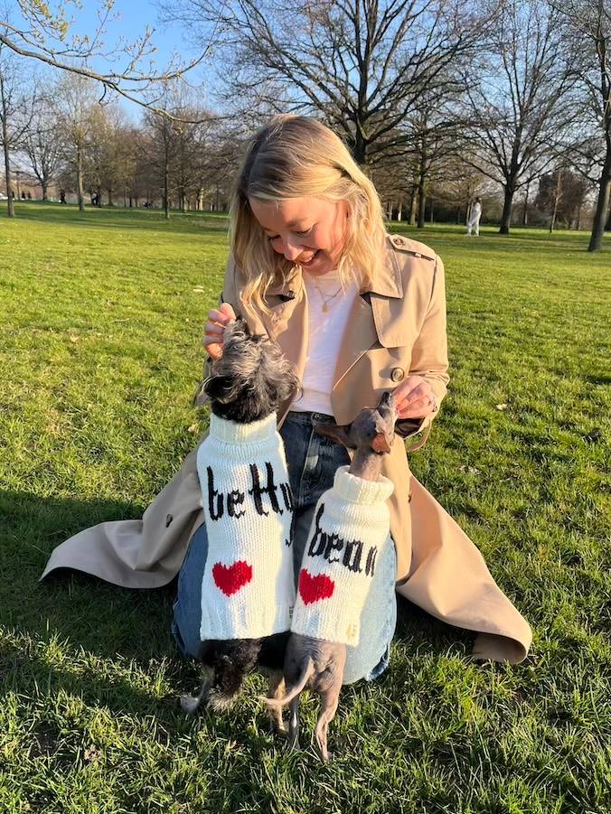 a woman with blonde hair sits in a park with two little dogs in sweaters jumping up on her