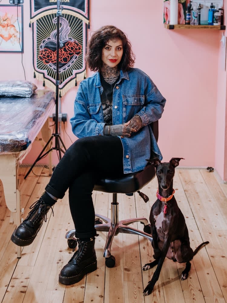 a woman with tattoos sits next to a black whippet in a tattoo studio