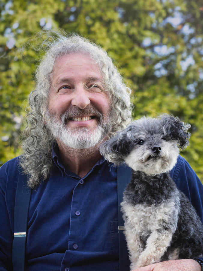 A man with long grey hair and a grey beard posing next to his small grey and white-haired dog who looks very similar.