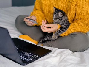 A woman in a bright yellow-orange sweater holding a striped kitten in one hand and a credit card in the other while using her laptop in front of her