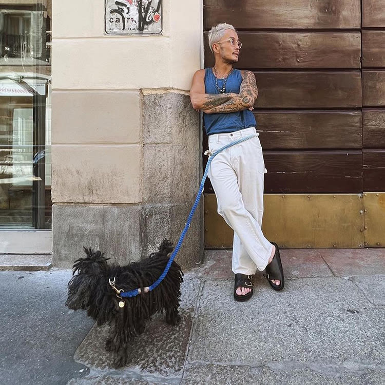 Person with white blond hair and tattoos wearing a blue muscle t-shirt and white pants and a hands-free blue leash connected to a black shaggy haired dog