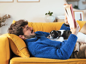 Man reading a book at home with small dog laying on his stomach.