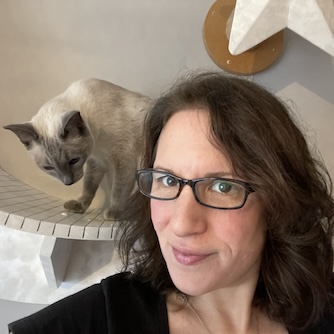 a woman with brown hair and glasses poses with a siamese cat behind her