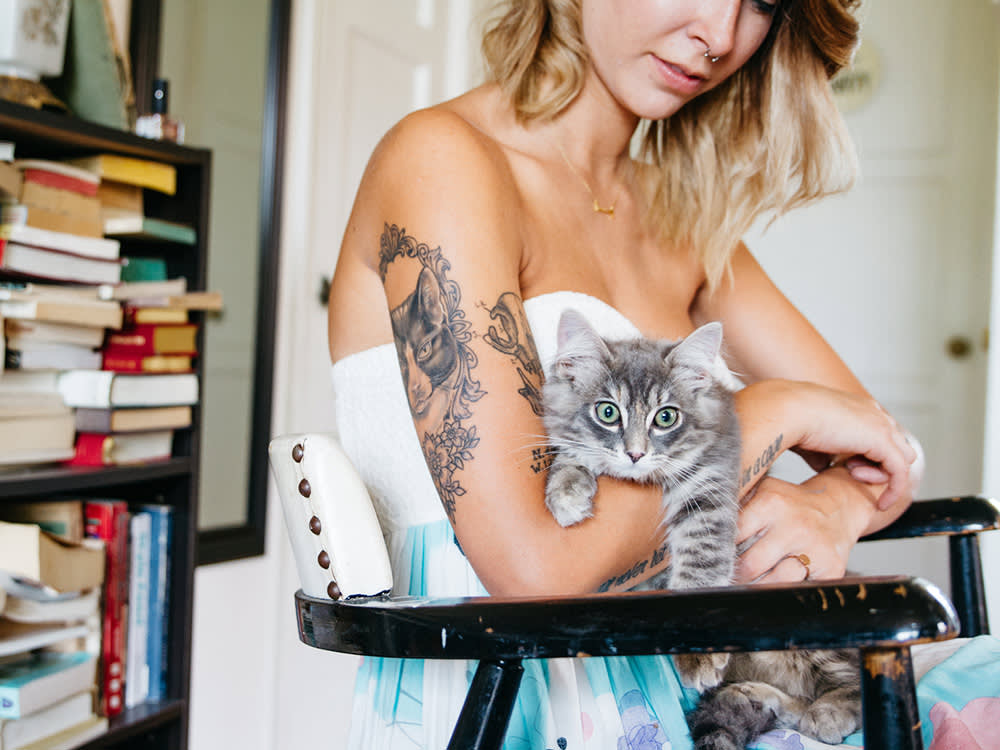 Young Woman With Tattoos Holds Cat In Home While Sitting In Chair.
