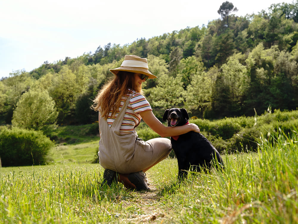 A woman in beige overalls a striped shirt and a straw hat, smiling, with her arm around her black dog sitting in the grass on a green hill.